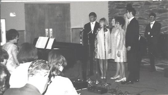 Vocalists sing harmoniously while a pianist accompanies them in a cozy venue.