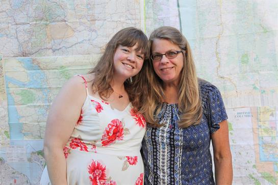 Two women stand together, smiling widely, in front of a map backdrop at a bright summer gathering.