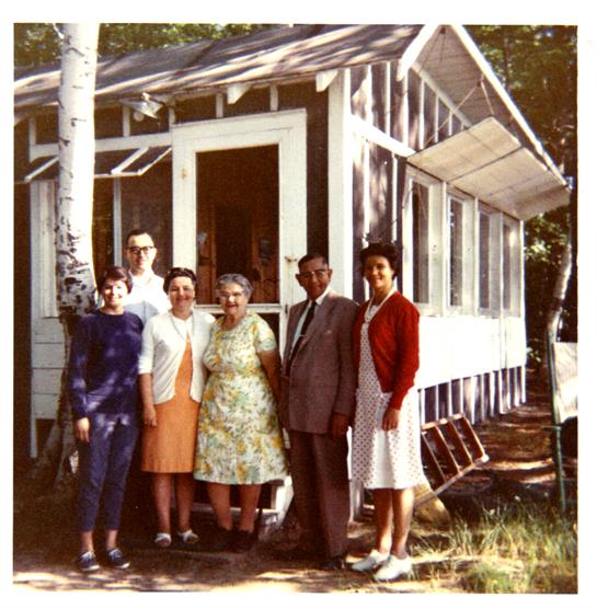 Several individuals pose together outside a modest cabin surrounded by trees on a sunny day.