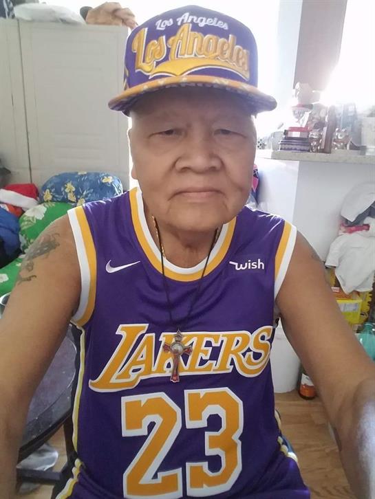 An elderly man proudly displays his Lakers attire, sitting amidst a lively, cluttered room.