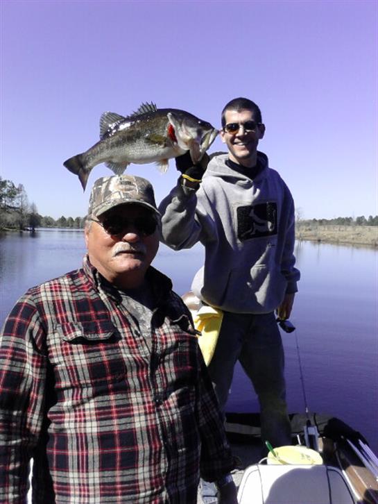 Two men are enjoying a fishing trip, showcasing a large bass caught on a sunny afternoon.
