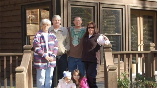 Group of six family members posing happily outside a cozy wooden cabin in warm weather.