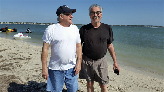Two men stand by the water's edge, laughing together under a clear blue sky on a beach.