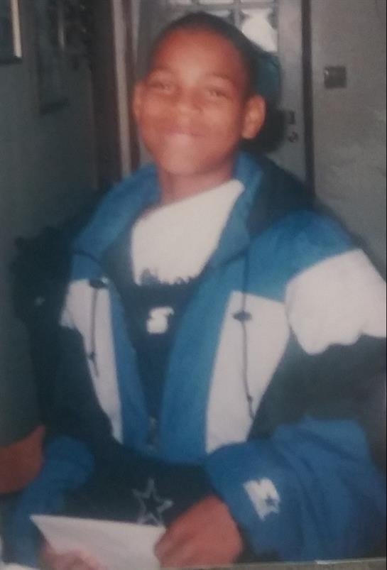 Young boy smiles while sitting indoors, dressed in a colorful jacket during sunset hours.