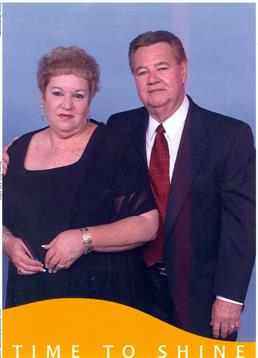 A well-dressed couple stands together in a studio, showcasing their attire and smiles.