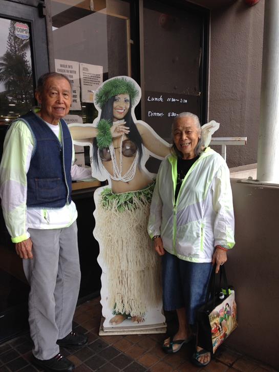 Two elderly men stand smiling beside a hula girl cardboard cutout at a cafe in Hawaii.