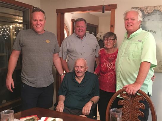 Several family members gather around an elderly man celebrating a milestone birthday in a home.