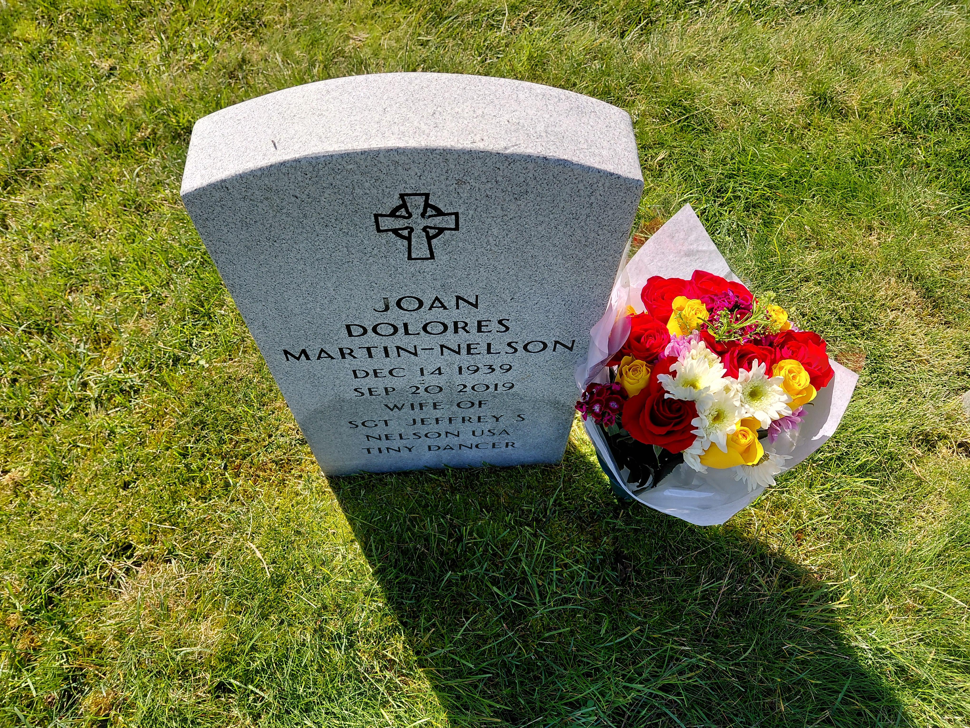 A gravestone marks the resting place of Joan Martin Nelson, surrounded by colorful flowers.
