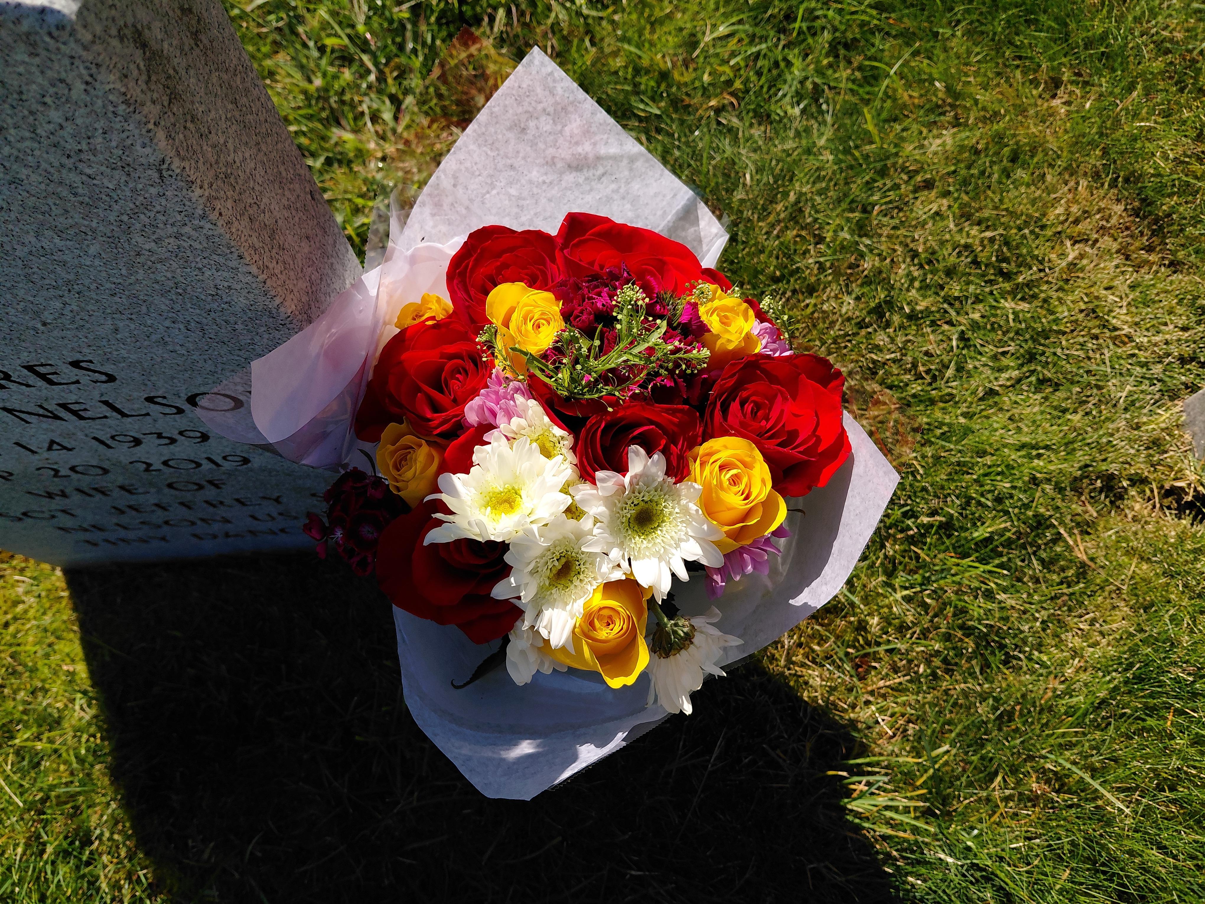 A vibrant bouquet with red, yellow, and white flowers sits on a grave in a peaceful cemetery.