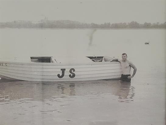 A man enjoys a relaxed moment in shallow water next to a boat on a calm lake surrounded by nature.