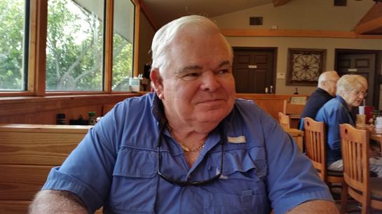 A man with white hair smiles while sitting at a table in a restaurant surrounded by warm sunlight.