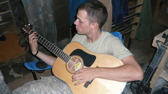 A young man strums an acoustic guitar, enjoying music in a casual military environment.