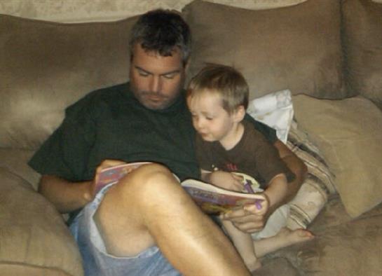 A father sits on a couch with his young son, engrossed in reading a storybook together.