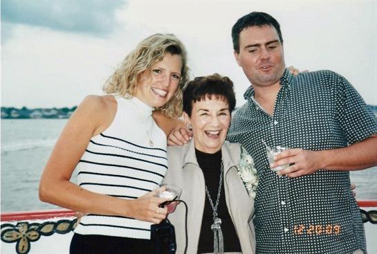 Three people happily celebrate outdoors by the water, posing with drinks.