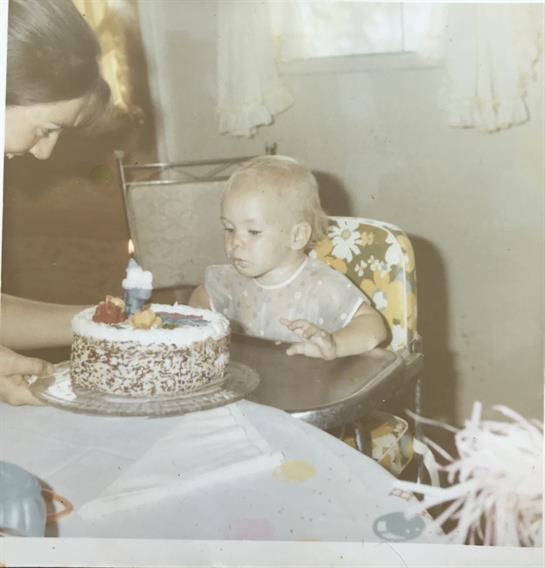 A young child in a high chair gazes at a lit birthday cake, surrounded by family.