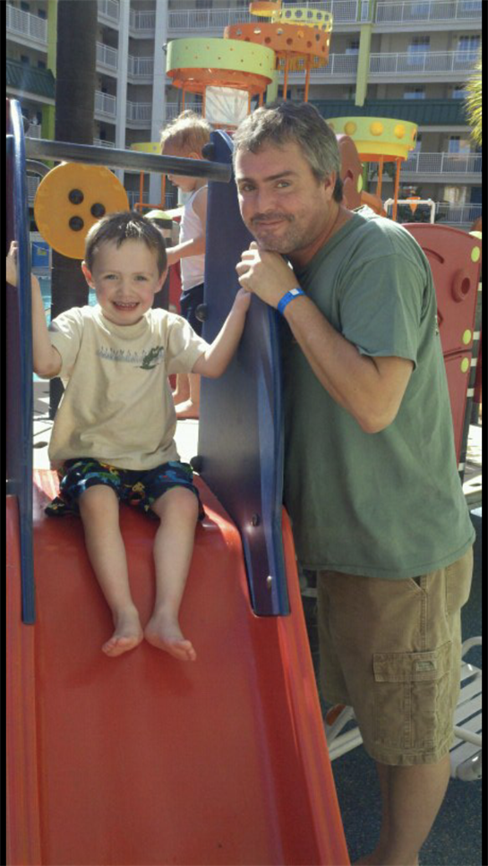 Child slides down with joy while father watches and smiles at a vibrant playground.