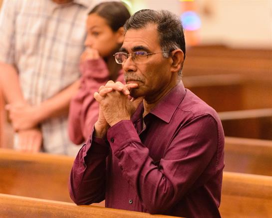 A man with glasses prays thoughtfully in a wooden pew during a gathering.