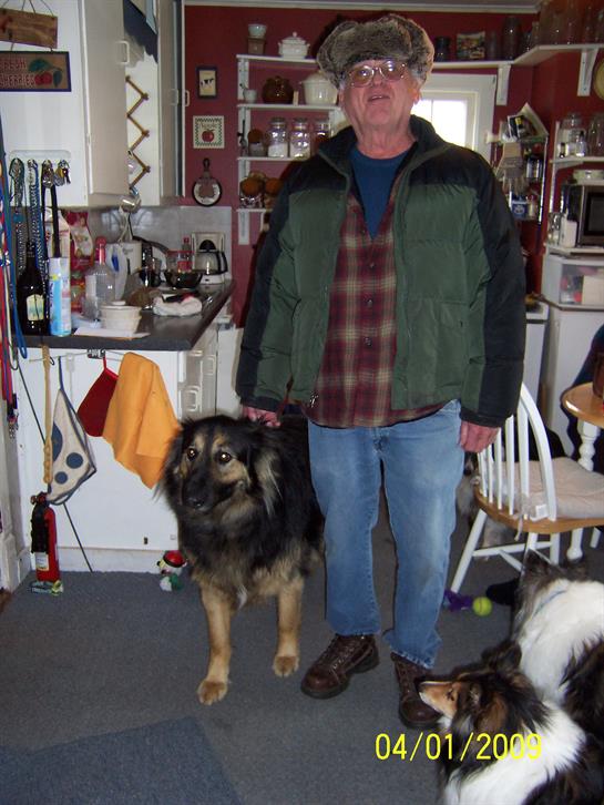 A man dressed in casual clothes stands in a warm kitchen accompanied by two friendly dogs.