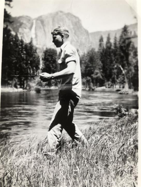 A man stands by the river, casting his line into clear water with mountains behind.