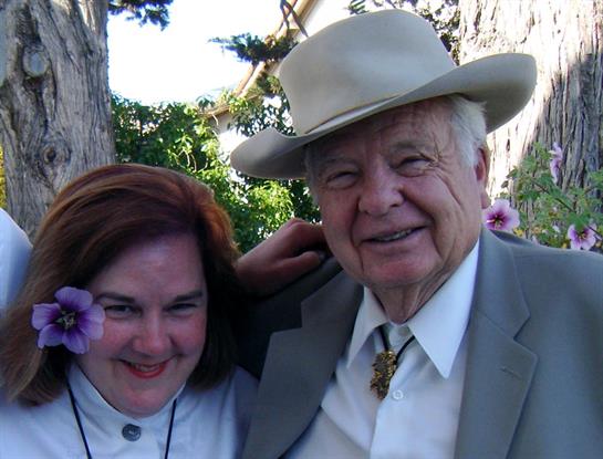 A man and woman stand close together, smiling brightly in a vibrant garden filled with flowers.