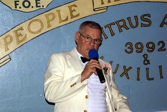 A man dressed in a white tuxedo speaks into a microphone at an event, engaging the audience.