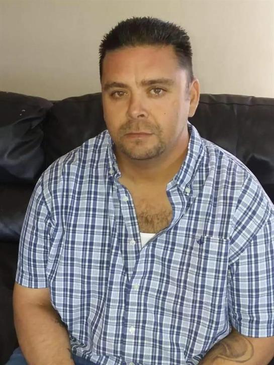 A man sits on a couch in a plaid shirt, appearing thoughtful while relaxing indoors.