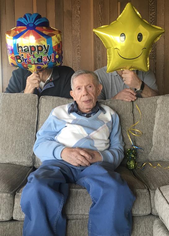 An elderly man enjoys a birthday celebration surrounded by family holding balloons.