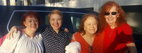 Two joyful women stand close, sharing smiles and warmth during a bright day outdoors.