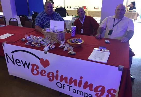 Three men seated at a table share information and refreshments at a local community event in Tampa.