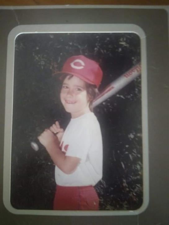 A child smiles while holding a baseball bat, wearing a red cap and a white shirt in the backyard.
