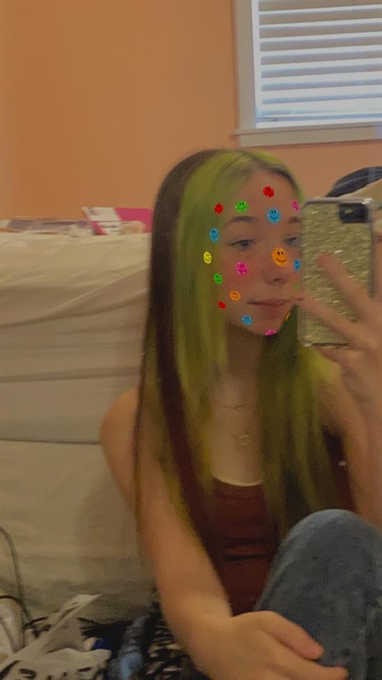 Girl with vibrant green hair and playful stickers on her face is capturing a selfie indoors.