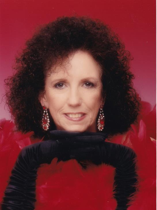 Woman with curly hair wears bright outfit and earrings, posing confidently against a red backdrop.