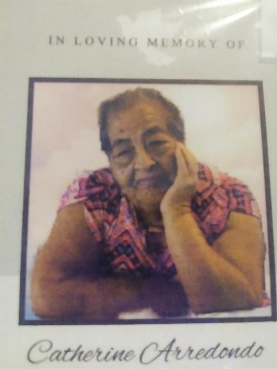 An elderly woman rests her chin on her hand while sitting indoors. She has a thoughtful expression.