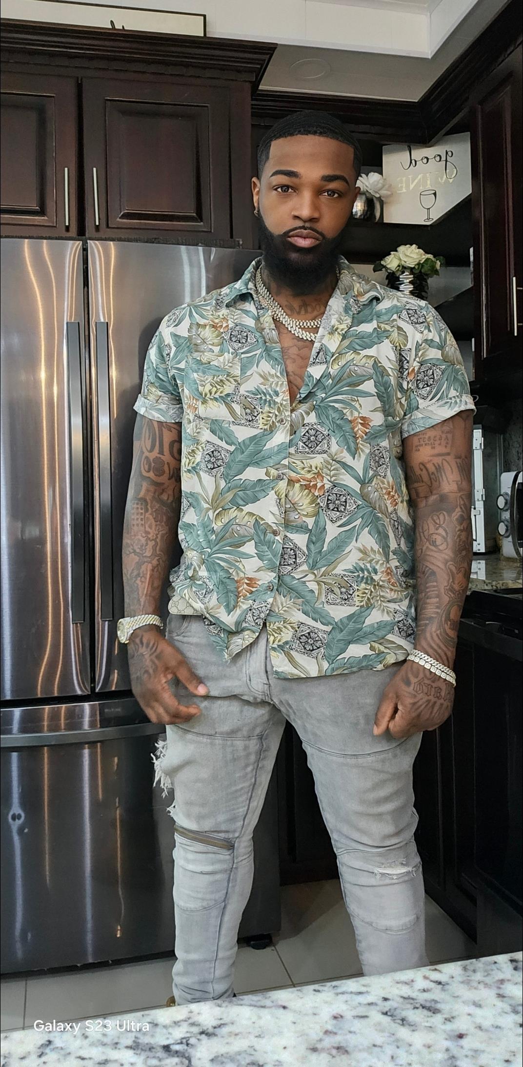 Man in a floral shirt and ripped jeans flaunts tattoos and confidence in a modern kitchen.