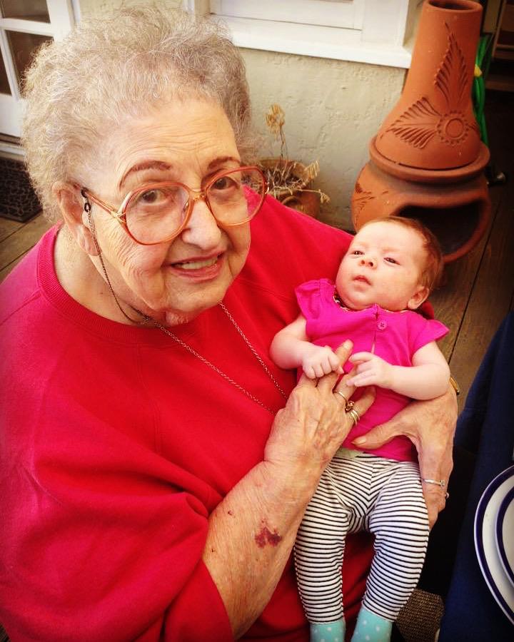 An elderly woman smiles warmly while cradling her baby granddaughter in a sunny backyard.