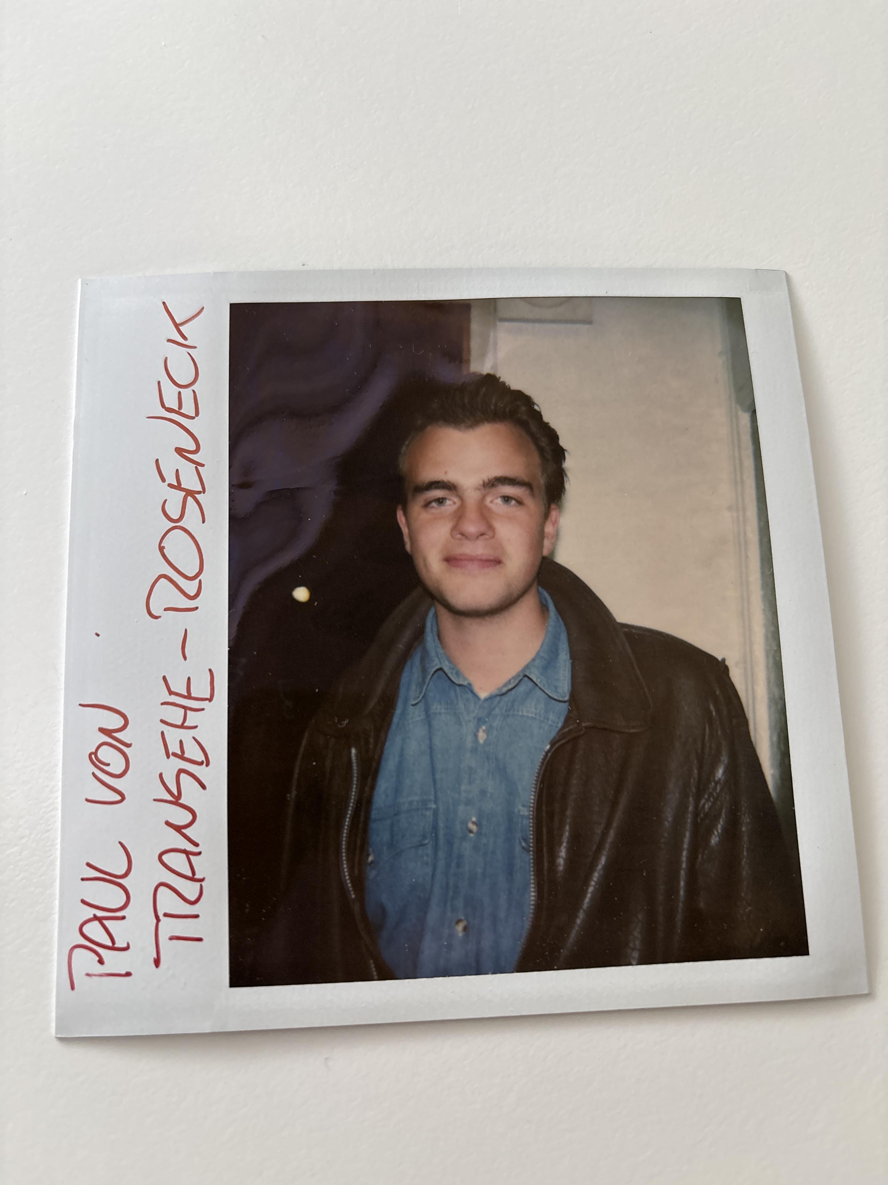 Young man stands confidently, wearing a leather jacket and denim shirt, captured in indoor lighting.