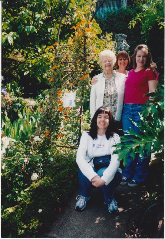 Four women pose happily in a beautifully landscaped garden filled with colorful flowers.