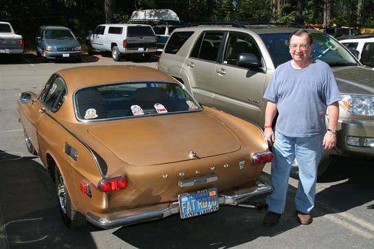 Man stands proudly by a classic brown car in a busy parking lot on a sunny afternoon.