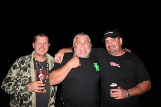 Three men are smiling and holding drinks together at a lively nighttime gathering.