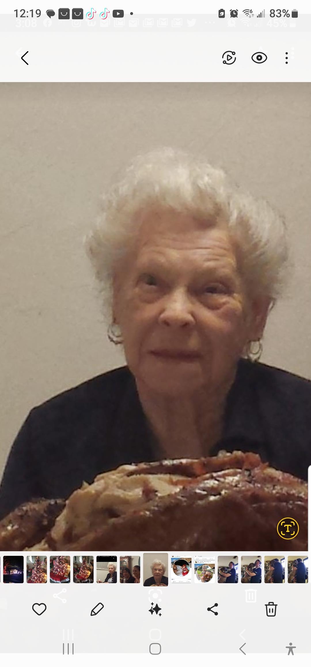 An elderly woman smiles softly while presenting a large dish during a family celebration.