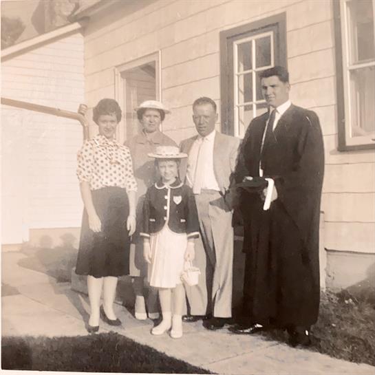A group of five people, dressed in formal attire, stand outside a house on a sunny day.