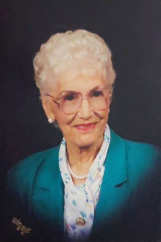 A smiling elderly woman with curly white hair is dressed in a turquoise blazer, looking confident.