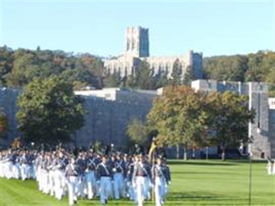 Group of cadets in uniform marching across a green field at a military academy during a sunny day.