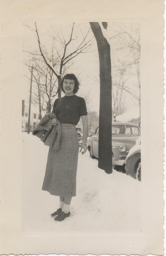 A woman confidently walks through deep snow in stylish vintage attire on a winter afternoon.