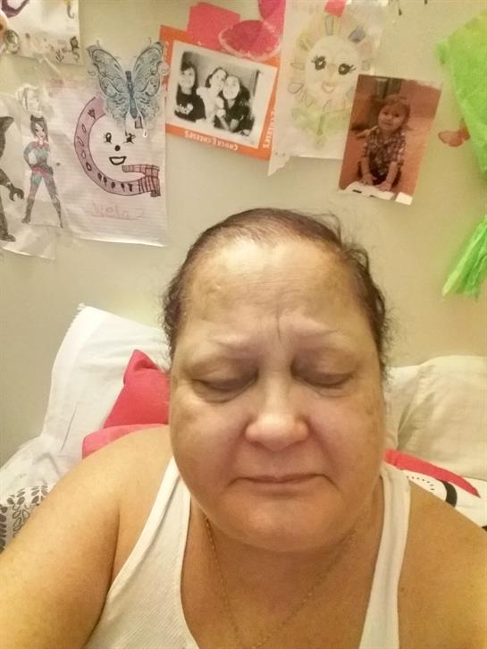 An elderly woman sits quietly on a bed, eyes closed in a cozy, decorated room filled with artwork.