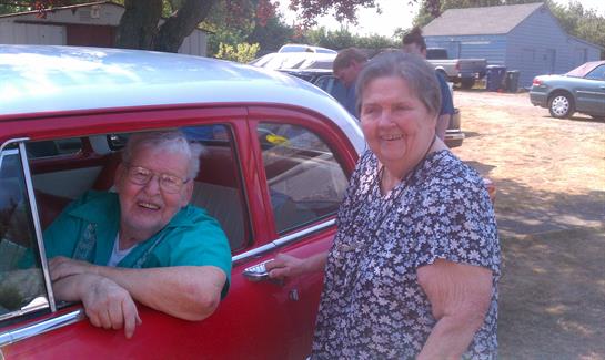 Two joyful women pose together near a vintage red car, enjoying a sunny day outdoors.
