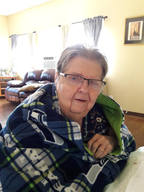 An elderly woman sits comfortably in her living room, wrapped in a warm blanket, smiling.