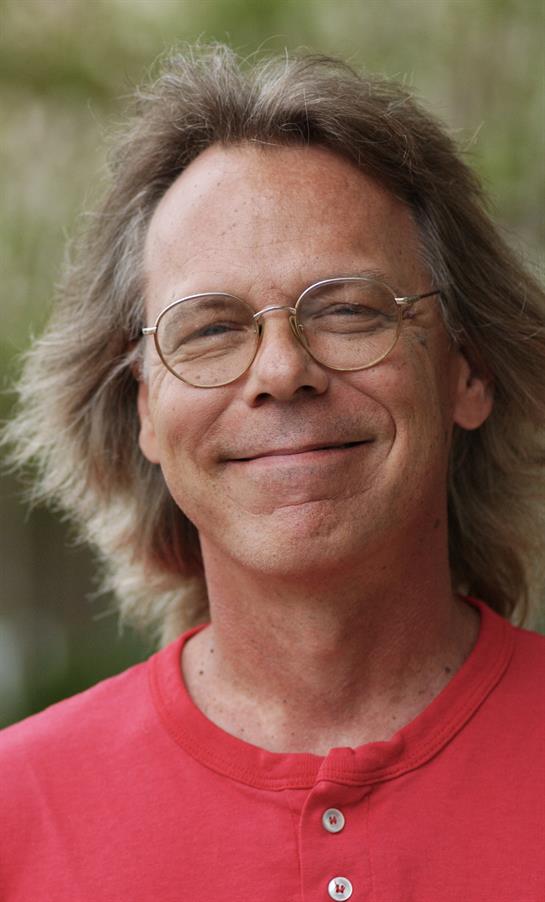 A man with long hair and glasses smiles warmly while enjoying the outdoors on a sunny day.