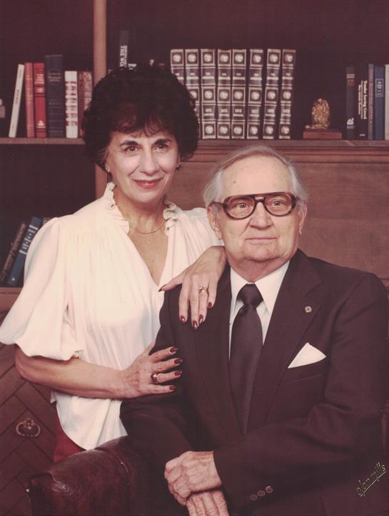 Elderly couple stands close together, smiling in a library filled with books and warm wood accents.