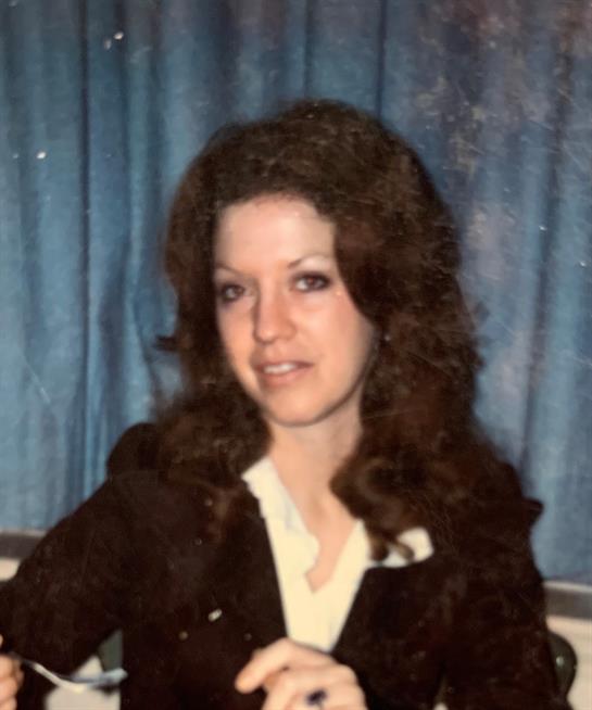 A woman with curly hair smiles in front of a blue curtain, displaying her vintage style.